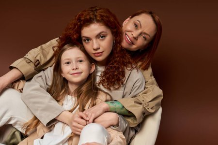tres generaciones, familia pelirroja positiva mirando a la cámara en el fondo marrón, crianza moderna