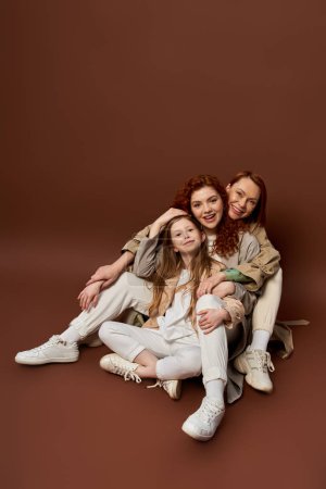 drei Generationen, fröhliche rothaarige Familie mit Sommersprossen, die vor braunem Hintergrund in die Kamera blickt