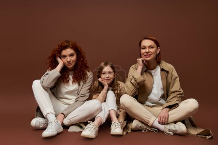 Téléchargez les photos : Heureux rousse famille de trois générations féminines assis sur fond brun, manteaux d'automne - en image libre de droit