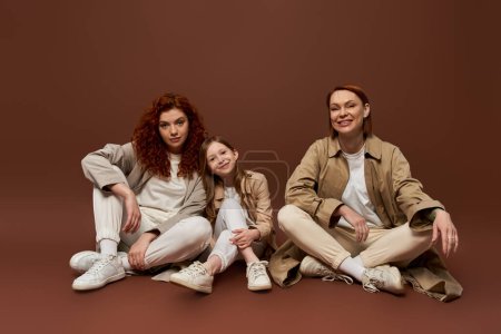 joyful redhead family of three female generations sitting on brown background, autumn fashion
