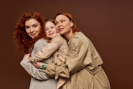 joyful redhead family of three female generations hugging on brown background, autumn fashion