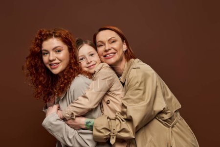 Téléchargez les photos : Heureux rousse famille de trois femmes générations étreignant sur fond brun, la mode d'automne - en image libre de droit