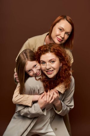 alegre familia pelirroja de tres generaciones femeninas en abrigos abrazándose unos a otros sobre fondo marrón