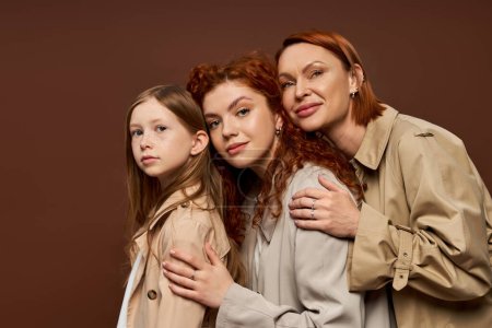 famille de trois générations féminines aux cheveux roux posant en manteaux beiges, étreignant sur fond brun