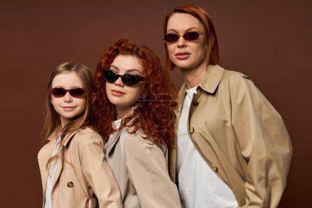 drei weibliche Generationen mit roten Haaren posieren mit Sonnenbrille vor braunem Hintergrund, die Hände in den Taschen