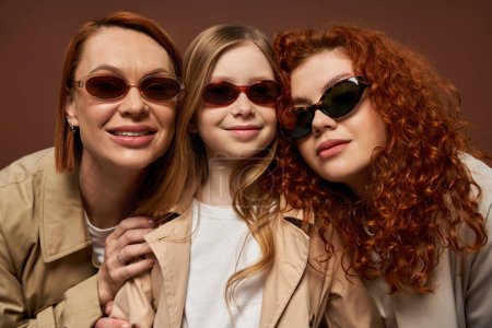 concept de changement de génération familiale, heureux rousses femmes et fille en lunettes de soleil sur fond brun