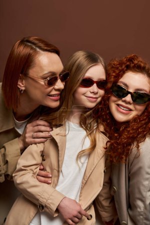 Konzept des Generationenwechsels, fröhliche rothaarige Frauen und Mädchen mit Sonnenbrille vor braunem Hintergrund