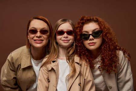 portrait of happy three generations of women looking at camera in sunglasses on brown backdrop