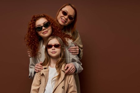 Foto de Retrato familiar de tres generaciones femeninas en gafas de sol y abrigos posando sobre fondo marrón - Imagen libre de derechos