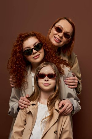 Foto de Retrato familiar de tres generaciones femeninas en gafas de sol y abrigos sonriendo sobre fondo marrón - Imagen libre de derechos