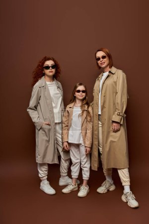 family photo of female generations in sunglasses and coats posing on brown background, full length