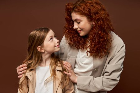 zwei Generationen, junge lockige Mutter mit roten Haaren, die Tochter auf braunem Hintergrund anschaut, Familie