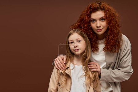 zwei Generationen, junge lockige Mutter mit roten Haaren, die mit ihrer Tochter vor braunem Hintergrund in die Kamera schaut