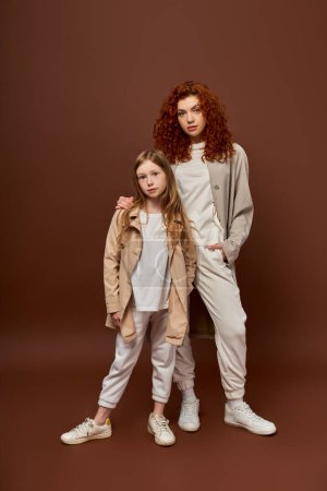 two generations, young curly mother and daughter looking at camera on brown backdrop, hand in pocket