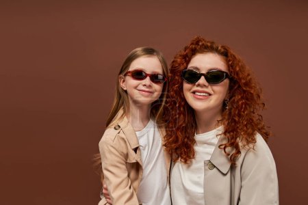two generations, happy curly mother and daughter in sunglasses on brown backdrop, portrait