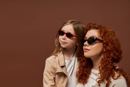 deux générations, heureuse rousse mère et fille en lunettes de soleil regardant loin sur fond brun
