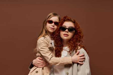 two generations, redhead mother and daughter in sunglasses and beige trench coats on brown backdrop