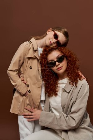 two generations, redhead mother and child in sunglasses and beige trench coats on brown backdrop