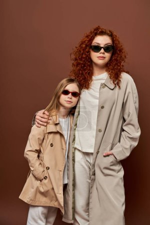 autumn style, redhead mother and kid posing in sunglasses and beige trench coats, brown backdrop
