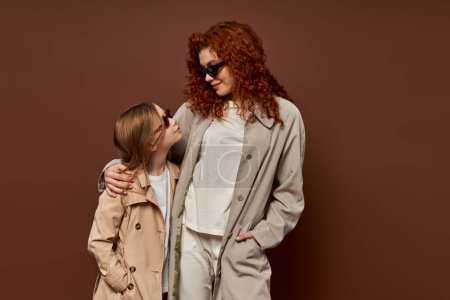 autumn fashion, redhead mother and kid posing in sunglasses and beige trench coats, brown backdrop