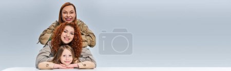 three generations, redhead mother and child in beige trench coats posing on grey background, banner
