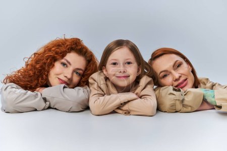 glückliche weibliche Generationen, rothaarige Frauen und Kind in beigen Mänteln lächelnd auf grauem Hintergrund, Familie