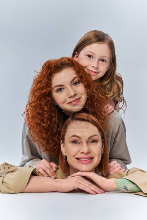 drei weibliche Generationen, glückliche rothaarige Familie in beigen Mänteln, lächelnd vor grauem Hintergrund, Familienbande