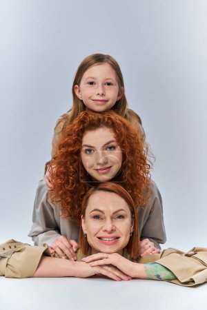 Foto de Tres generaciones, feliz familia pelirroja en abrigos beige sonriendo sobre fondo gris, vínculo femenino - Imagen libre de derechos