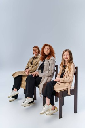 three female generations, happy redhead family in beige coats sitting on chairs on grey backdrop