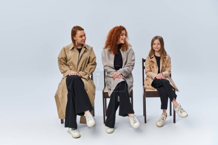 drei Generationen, rothaarige Frauen in Mänteln, die auf Stühlen sitzen und Mädchen vor grauem Hintergrund betrachten
