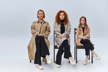 three female generations, redhead women and child in coats sitting on chairs on grey backdrop