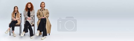 Photo for Three female generations, redhead women and kid in coats sitting on chairs on grey backdrop, banner - Royalty Free Image