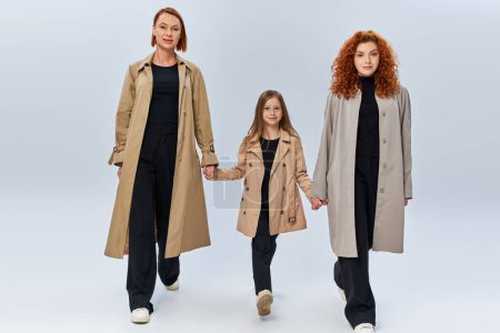 drei weibliche Generationen, rothaarige Frauen und Mädchen in Mänteln, Händchen haltend und vor grauem Hintergrund wandelnd