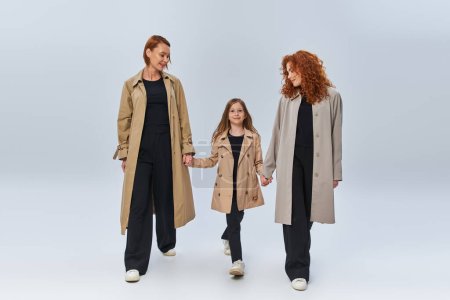 three generations, redhead women and girl in trench coats holding hands and walking on grey backdrop
