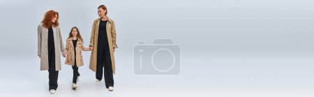 Téléchargez les photos : Trois générations, famille rousse en manteaux tenant la main et marchant sur fond gris, bannière - en image libre de droit