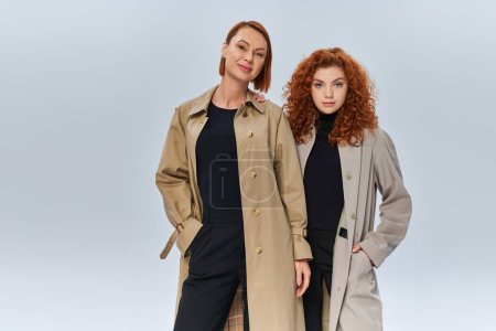 two generations of redhead women posing in coats and looking at camera on grey background, family