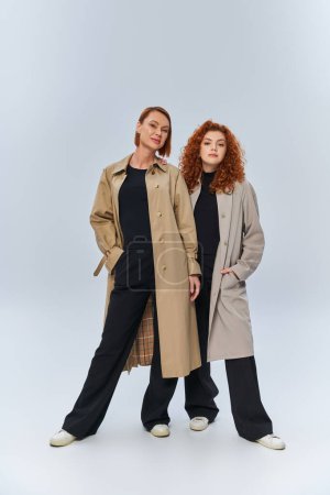 dos generaciones de mujeres pelirrojas posando en abrigos otoñales sobre fondo gris, manos en bolsillos