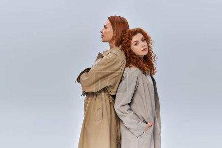 dos generaciones de mujeres pelirrojas de pie en abrigos de trinchera sobre fondo gris, moda otoñal