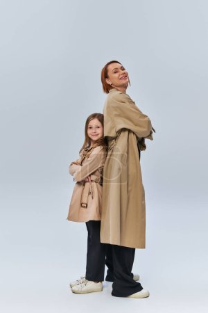 deux générations, heureuse rousse femme et fille debout dans des trench manteaux avec les bras croisés en studio