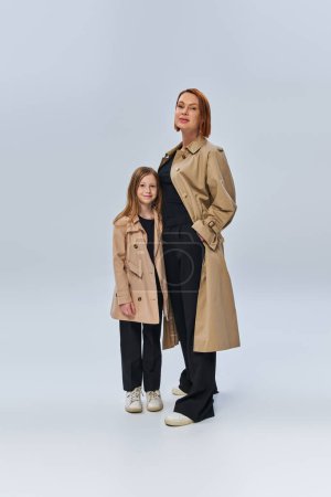 two female generations, redhead woman and happy girl standing in trench coats on grey background