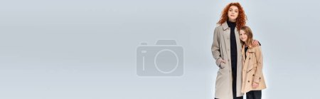 Téléchargez les photos : Deux générations, rousse femme embrassant fille et debout en manteaux sur fond gris, bannière - en image libre de droit