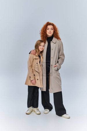 two female generations, redhead woman embracing happy kid and standing in coats on grey background