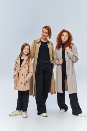 redhead family in trench coats posing together on grey background, three generations of women