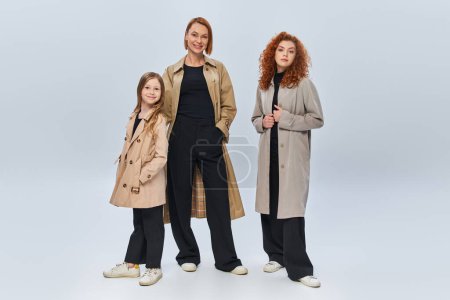 happy redhead family in trench coats posing together on grey background, three female generations