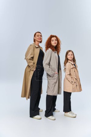 Foto de Alegre familia pelirroja en abrigos posando con las manos en bolsillos sobre fondo gris, generaciones femeninas - Imagen libre de derechos