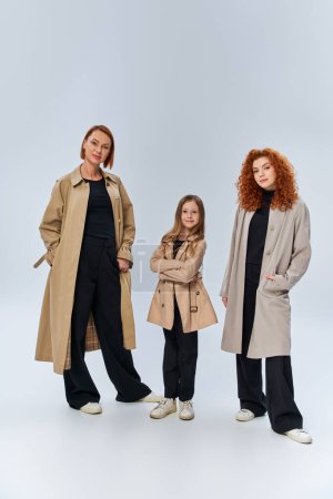familia pelirroja en abrigos posando con las manos en los bolsillos sobre fondo gris, generaciones femeninas felices
