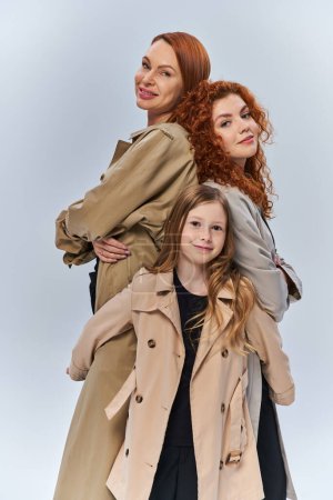 cute redhead girl hugging happy relatives in autumn coats on grey background, female generations