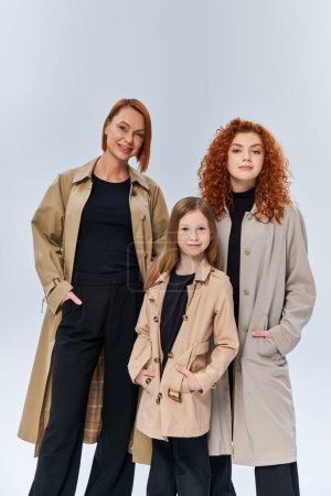 redhead girl posing near happy family in coats on grey backdrop, generations, hands in pockets