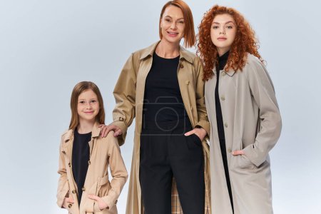 famille rousse en manteaux d'automne posant avec les mains dans les poches sur fond gris, générations féminines