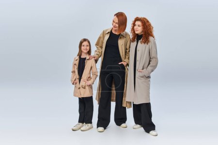 famille rousse en manteaux d'automne posant avec les mains dans les poches sur fond gris, trois générations
